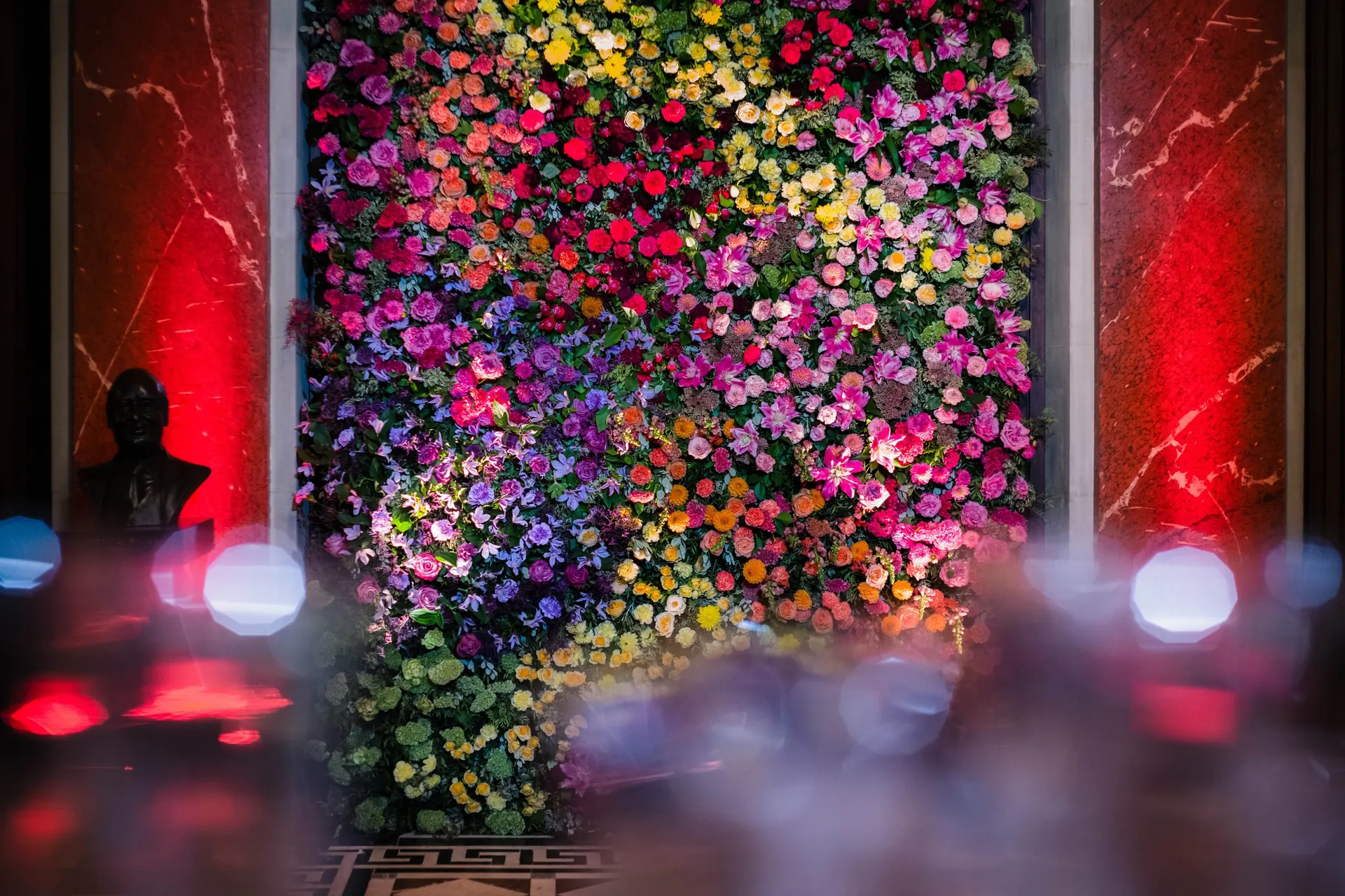 Un photocall en mur de fleurs multicolores, entre deux colonnes de marbre éclairées de rouge, dans le hall d’un théâtre